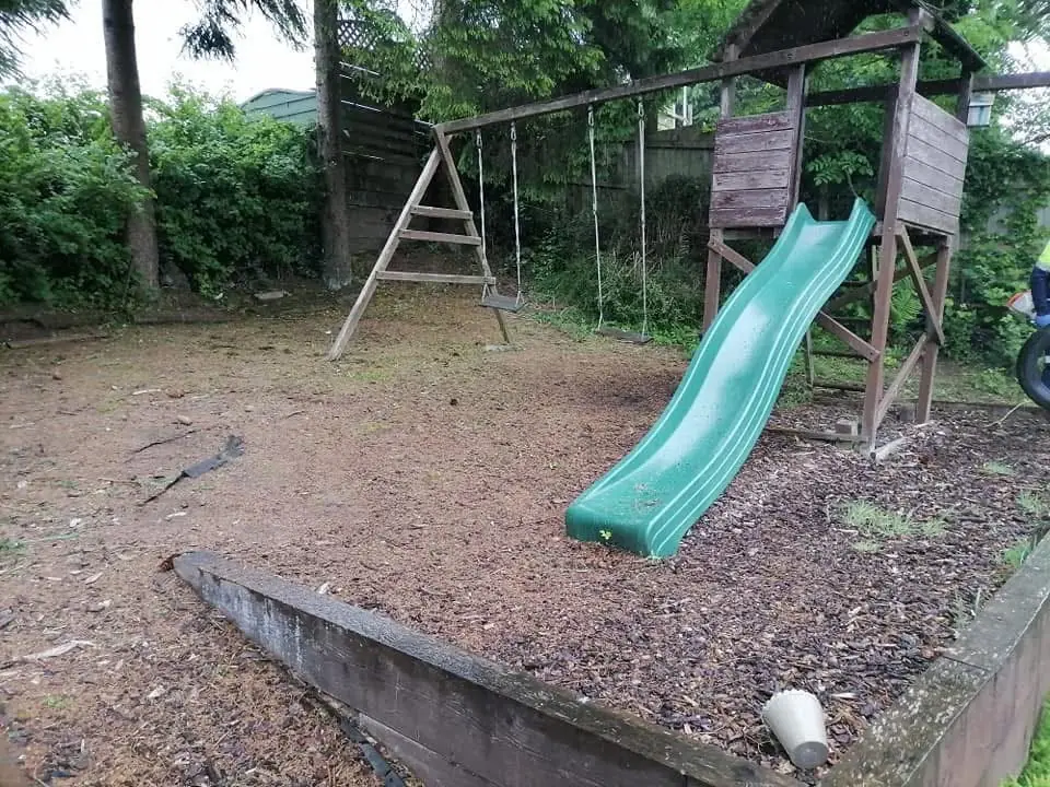 Bare, worn ground under a swing and slide frame, with a sleeper edge